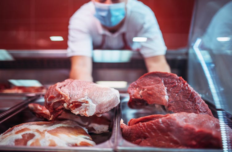 A person wearing a mask stands behind a counter of cuts of red meat