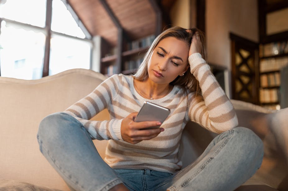 A woman in casual clothing sits on a sofa and looks concerned at her mobile phone
