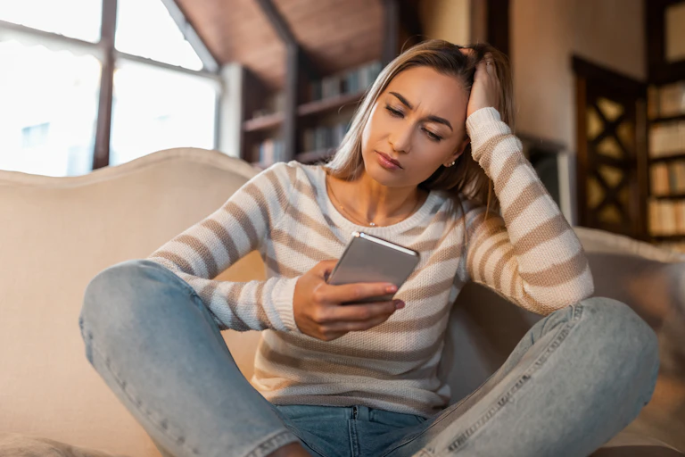 A woman in casual clothing sits on a sofa and looks concerned at her mobile phone