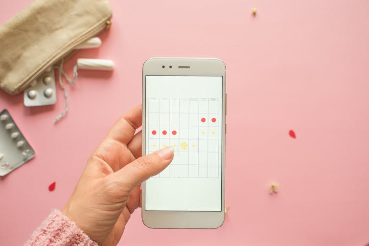 Overhead view of a woman's hand holding a mobile phone with a menstrual calendar app on screen. A few tampons and birth control pills are on the pink table underneath her phone.