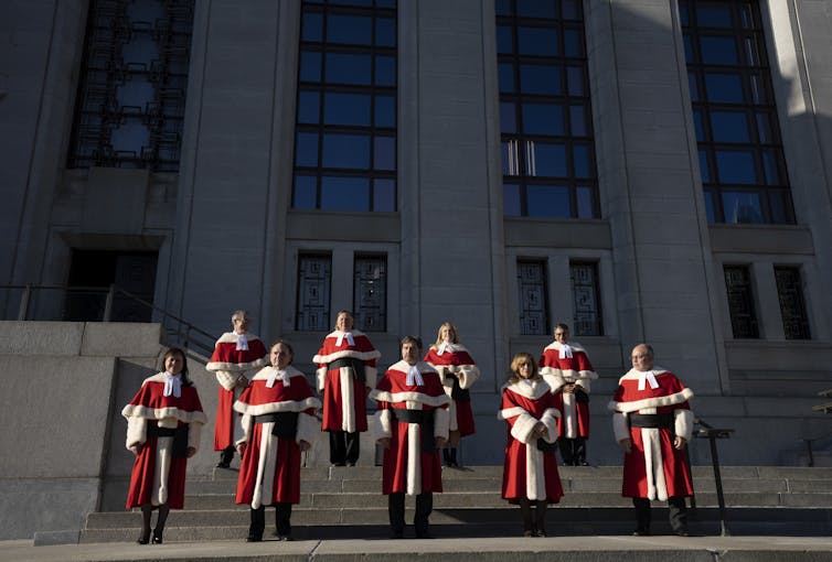 Nine judges in red and white robes stand outside the Supreme Court of Canada.