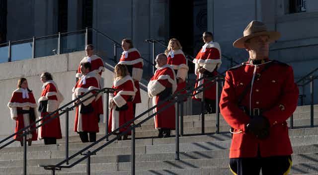 Nine people in red and white robes stand on the steps of the Supreme Court of Canada. A Mountie is in the foreground.