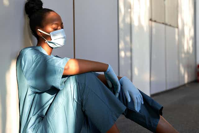 A woman wearing scrubs, gloves and a mask sits leaned up against a wall