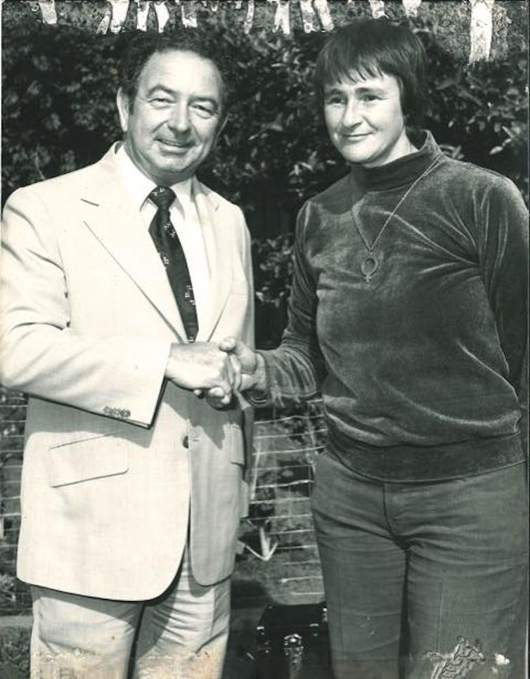 A man in a suit shakes the hand of a serious-looking woman with short hair.