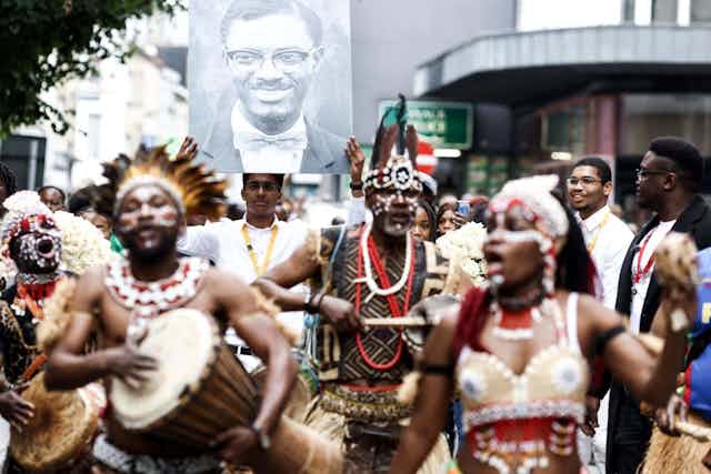 A crowd of people in the street drumming and chanting wearing traditional attire, a man in the background carrying a large poster of a photo of a man with glasses, smiling.
