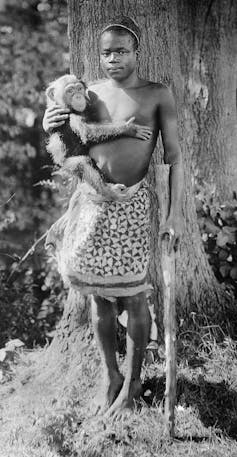 A young African man poses with a chimpanzee, holding on to a stick and wearing a traditional skirt