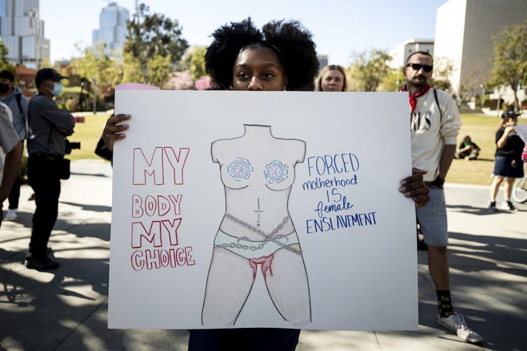 A woman holds a handmade sign at a rally, which features an illlustration of a headless woman's body in chains, and text reading 'my body my choice, forced motherhood is female enslavement'.
