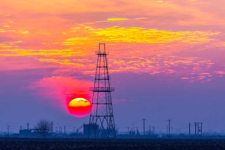 An oil and gas rig on land silhouetted against the setting sun.