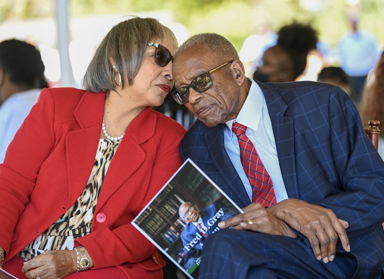 An older woman in a red jacket whispers into the ear of an older man in a blue suit and red tie.