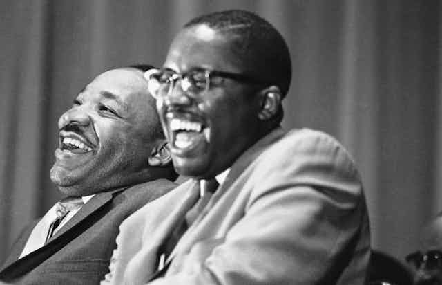 Two men in suits, sitting next to each other and laughing.
