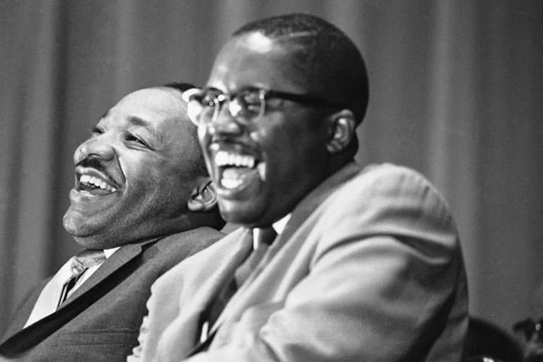 Two men in suits, sitting next to each other and laughing.