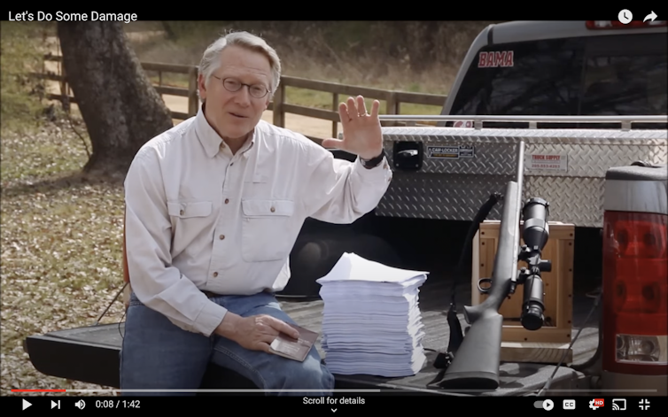 A middle-aged white man sits in the back of a pickup truck with a stack of papers and a high-powered rifle.