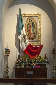 An illustration of the Virgin Mary inside a gold frame hangs on a wall beside a Mexican flag.
