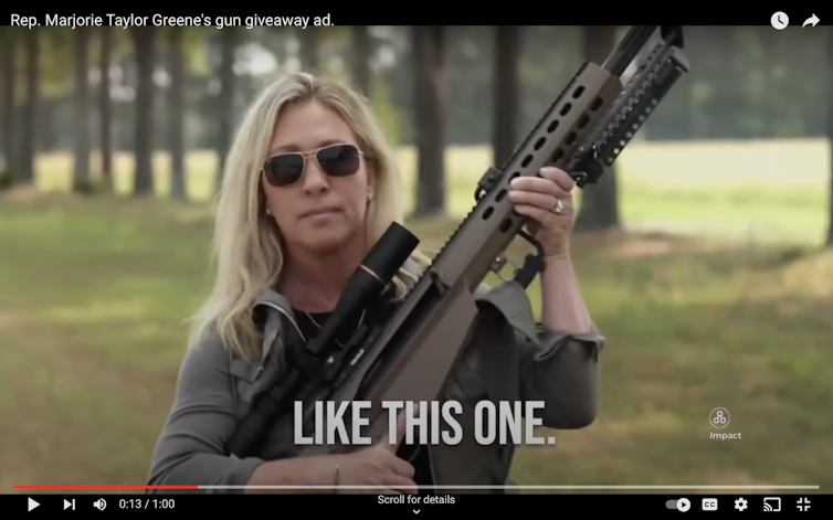 A white woman is wearing dark sunglasses and carrying a high-powered rifle.