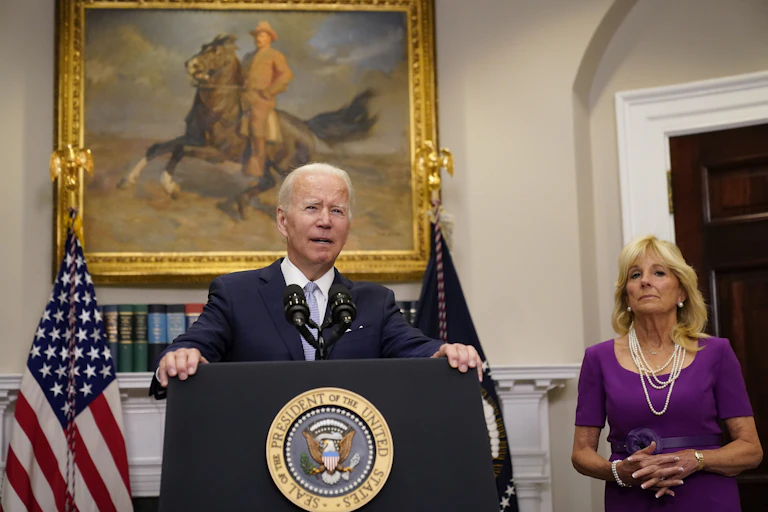 A man in a blue jacket and a tie, standing giving a speech in front of a painting of former President Teddy Roosevelt, with a bold woman wearing pearls standing to the side.