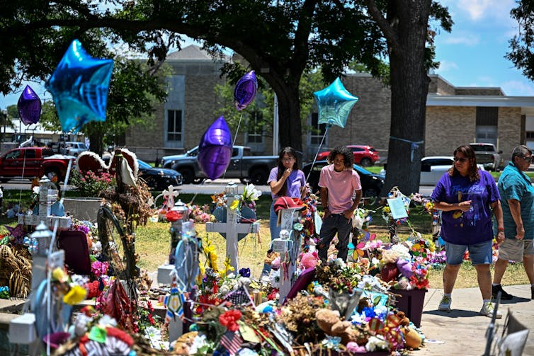Crosses, balloons, flowers, flags and other items in a memorial in front of a building.