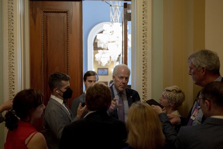 A white-haired man in a suit trying to make his way out of a room with high ceilings into a hallway filled with people crowding around him.
