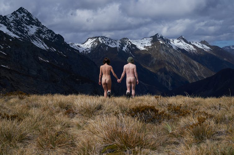 Two naked people hold hands walking away from the camera