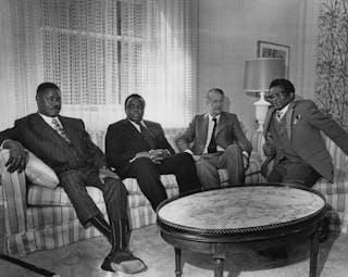 Four men in suits sitting on couches around a coffee table in a lounge setting.