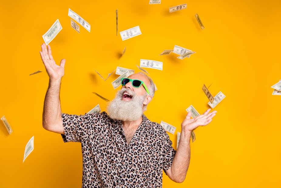 A bearded man in sunglasses and a leopard-print shirt smiles with his hands in the air as dollar bills rain down around him. Background is bright yellow.