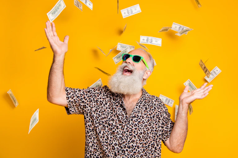 A bearded man in sunglasses and a leopard-print shirt smiles with his hands in the air as dollar bills rain down around him. Background is bright yellow.