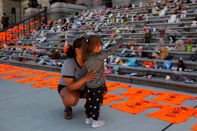 A woman holds her daughter in front of steps covered in toys and childrens shoes