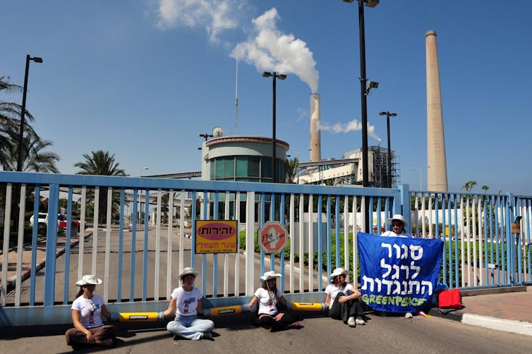 Five people sit outside a gate in front of a factory with steam billowing from a tall chimney