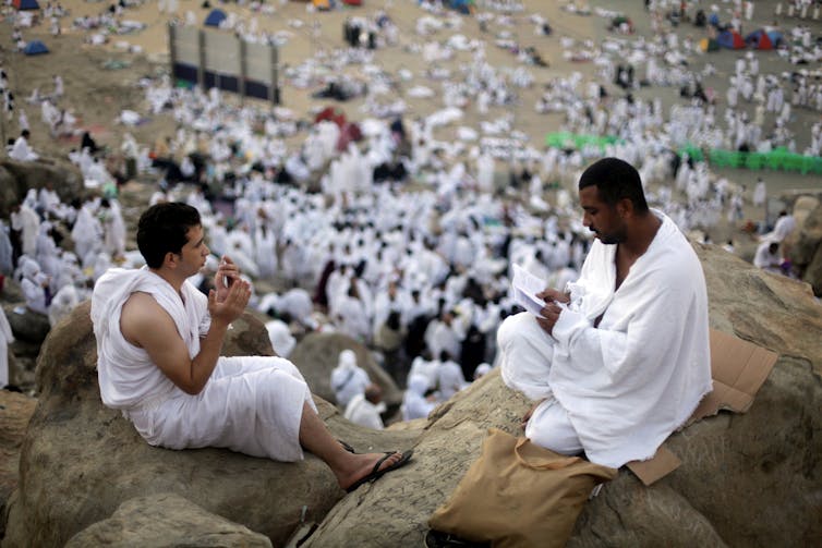 Two men dressed in loose white garments sitting on top of a hill, while multitudes of people are gathered below.