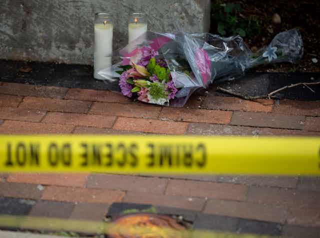 Flowers and candles are seen on a sidewalk behind yellow tape marking a crime scene.