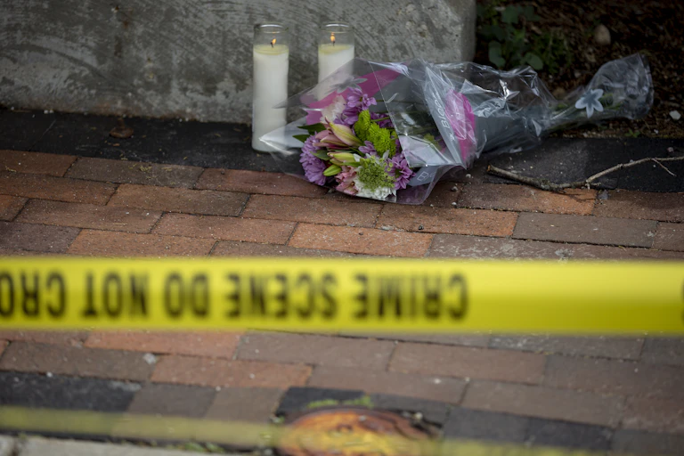 Flowers and candles are seen on a sidewalk behind yellow tape marking a crime scene.