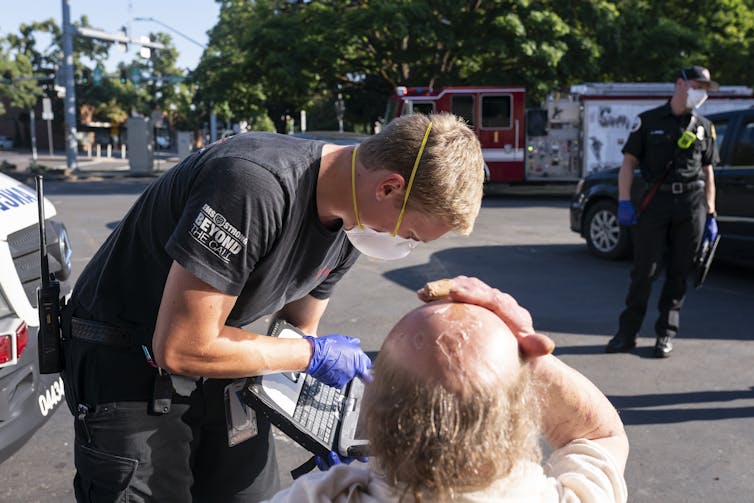 A paramedic in a mask bending down to talk to a seated man pressing a hand to his head. The paramedic is pointing to the screen of a small laptop he is holding.