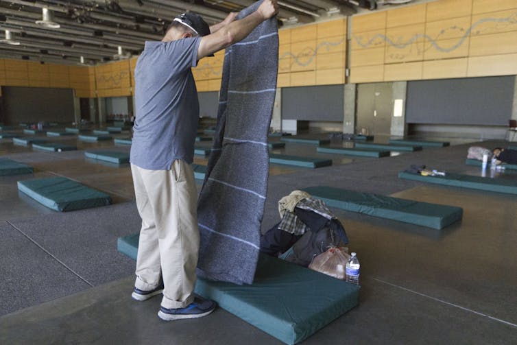 A man unfolds a blanket above a mattress lying on the floor of a large room. There are several identical mattresses lying on the floor nearby.