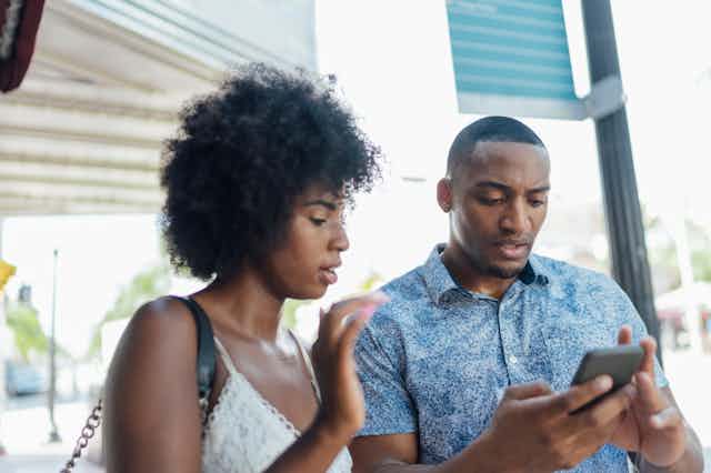 A young man holds a smart phone in both hands with a puzzled look on his face as a young woman looks at the screen and gestures with her right hand