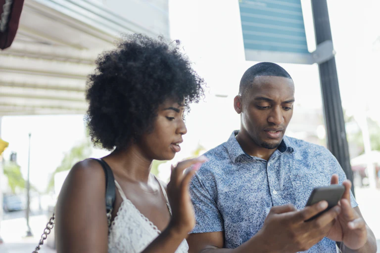 A young man holds a smart phone in both hands with a puzzled look on his face as a young woman looks at the screen and gestures with her right hand