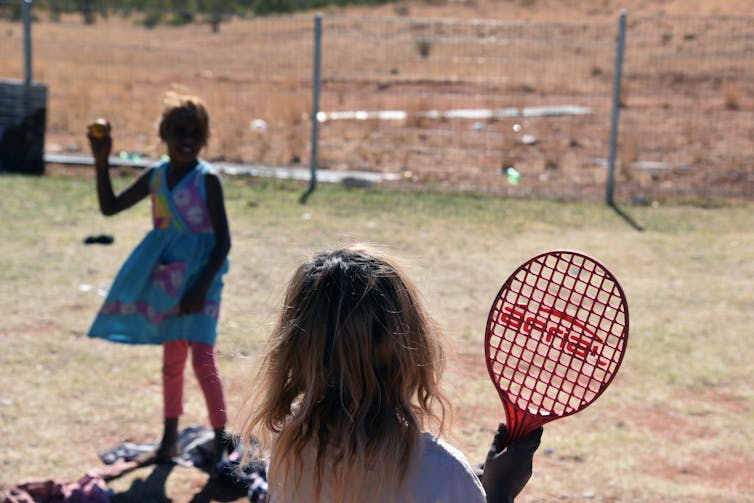 Two First Nations children play with a tennis racket and ball.