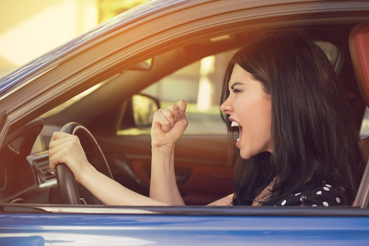 An angry women with dark hair in a driver's seat screams and raises a fist.