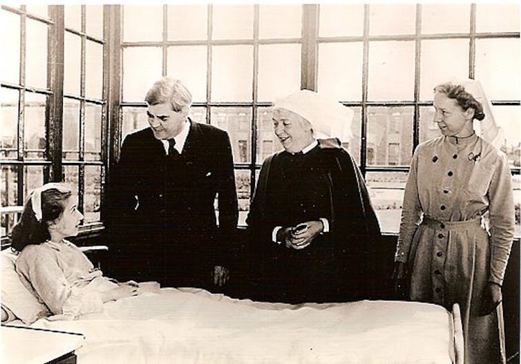 Three people stand around a patient's bed in an old hospital.
