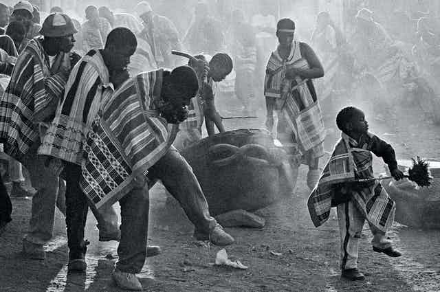 A black and white photo of a traditional African ceremony. Men in blankets dance behind a child holding feathers, drummers in the background and behind them members of the community.