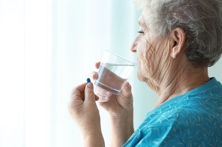 An elderly woman sips a glass of water. She is holding a blue pill between her fingers in her other hand.