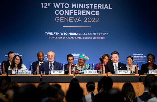 A panel of ministers are seated a table with a backdrop that reads 12th WTO Ministerial Conference Geneva 2022.