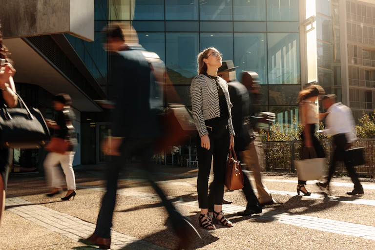 A woman stands still looking up at the sky in front of an office building, surrounded by blurred figures
