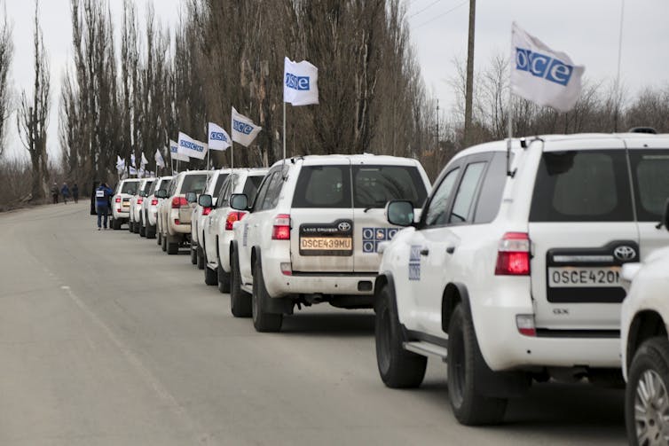 A column of white SUVs with blue and white flags line a road, ensuring European Security.