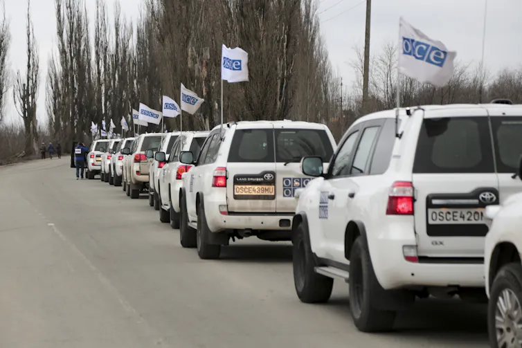 A column of white SUVs with blue and white flags line a road.