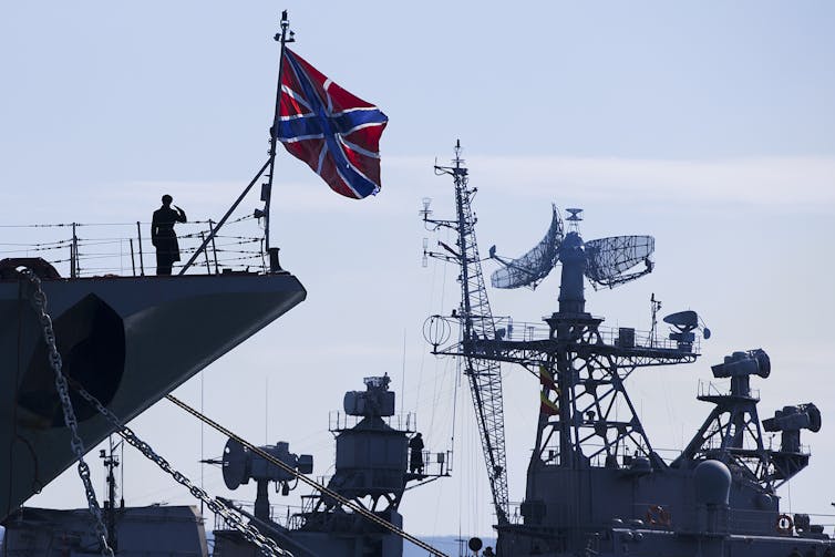 A sailor under the red-and-blue Russian flag salutes as another ship passes.