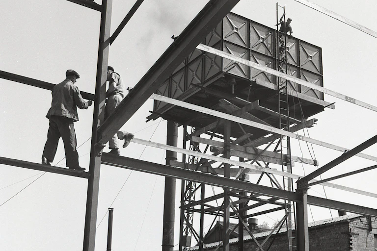 Two construction workers in suits stand on scaffolding beams.