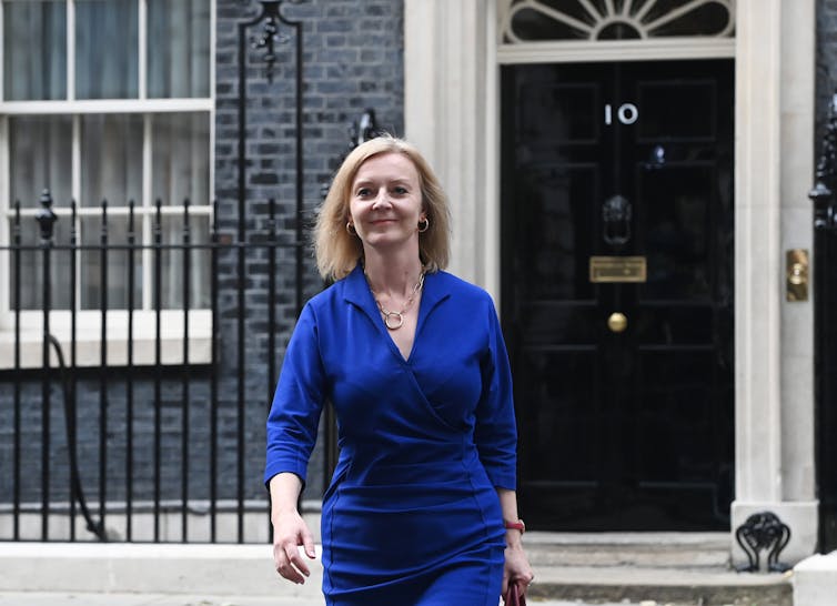 Liz Truss in a dark blue dress, walking towards the camera with Number 10 Downing Street behind her