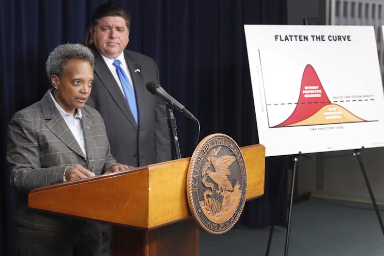 two people stand behind a podium, in the background a graph titled FLATTEN THE CURVE