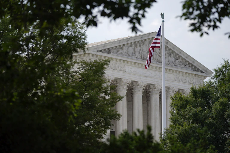 An American flag flutters outside the Supreme Court, frammed by green leafs of nearby trees.
