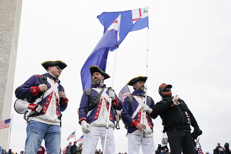 four men in colonial garb stand beneath a confederate flag