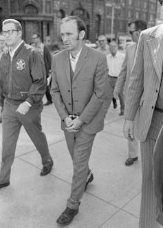 Man in suit walks with arms and legs handcuffed.